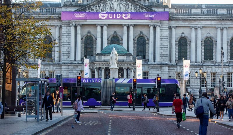 Glider bus passing by City Hall, Belfast as pedestrians cross the road in both directions.