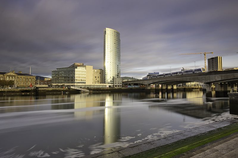 Train passing over Queen Elizabeth II Bridge in Belfast city centre, with city skyline in the background.